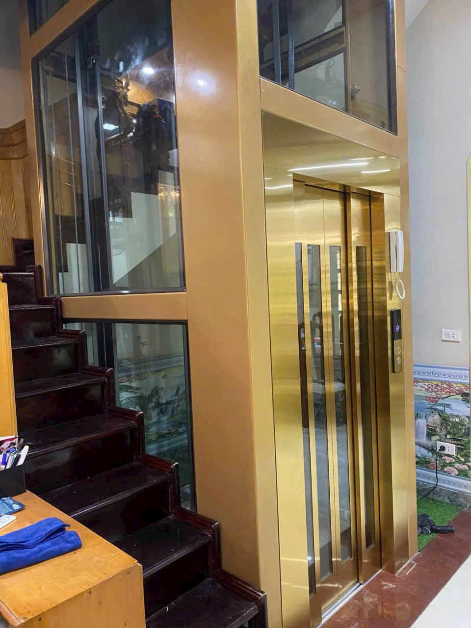 Thang máy gia đình vách kính khung vàng đặt cạnh cầu thang gỗ trong nhà